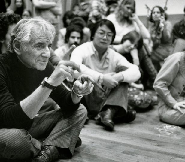 The archival photo (black and white) captures Leonard Bernstein in a candid moment during a master class. The maestro is seated on the floor, leaning forward attentively, surrounded by a group of focused students who are also sitting , reflecting the casual and intimate atmosphere of the learning experience.